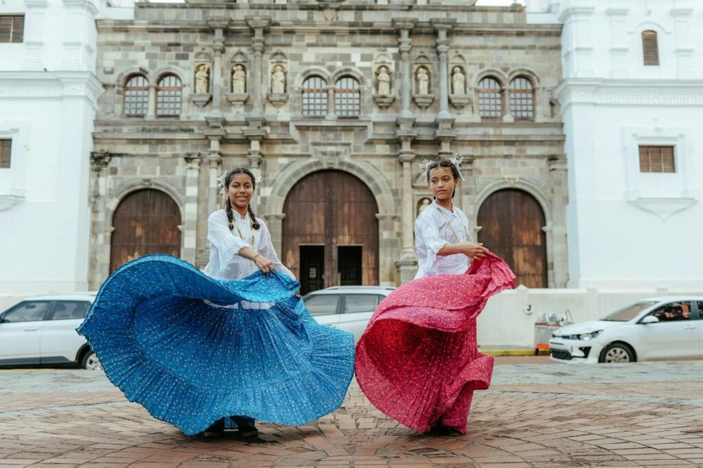 Two women in vibrant skirts perform a traditional dance in front of a historic building on a city street.